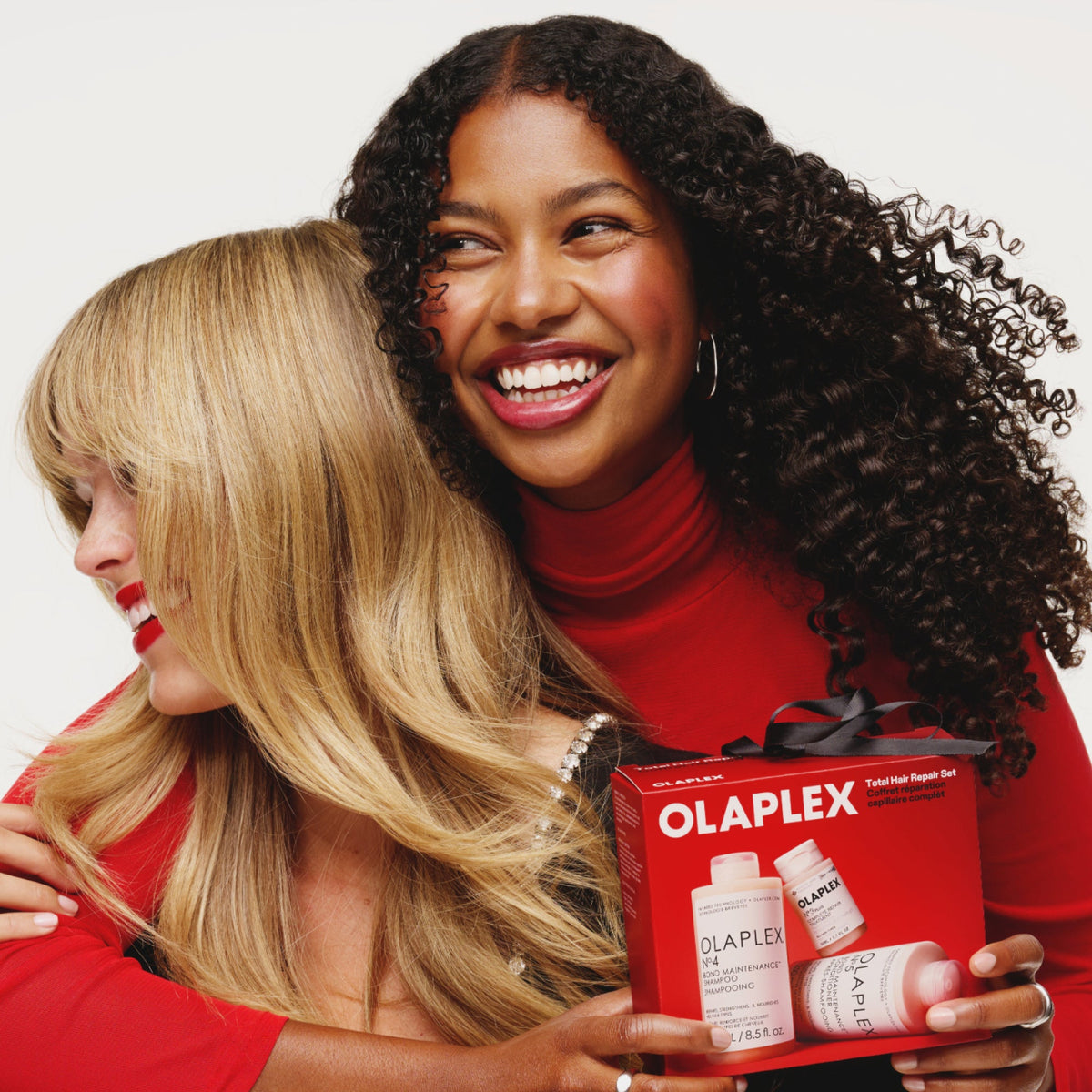 Two women with blonde and curly hair holding the OLAPLEX Total Hair Repair Set gift box